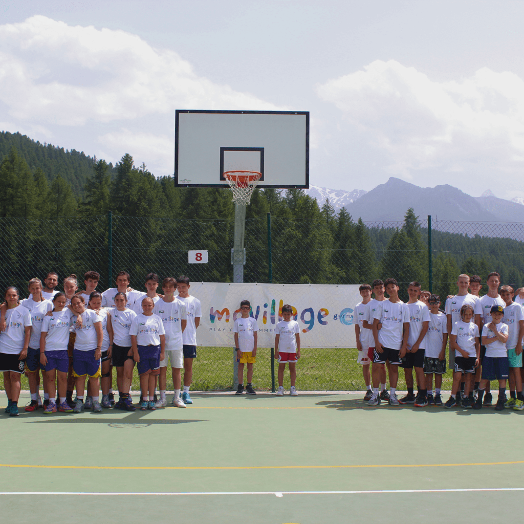 Foto di gruppo del Movillage Basket Camp: bambini e ragazzi sorridenti, indossano canotte da basket e tengono in mano il pallone, circondati dagli allenatori. Il gruppo è riunito su un campo da basket immerso nella natura, al termine di una giornata di allenamenti e giochi, in un clima di entusiasmo, amicizia e spirito di squadra.